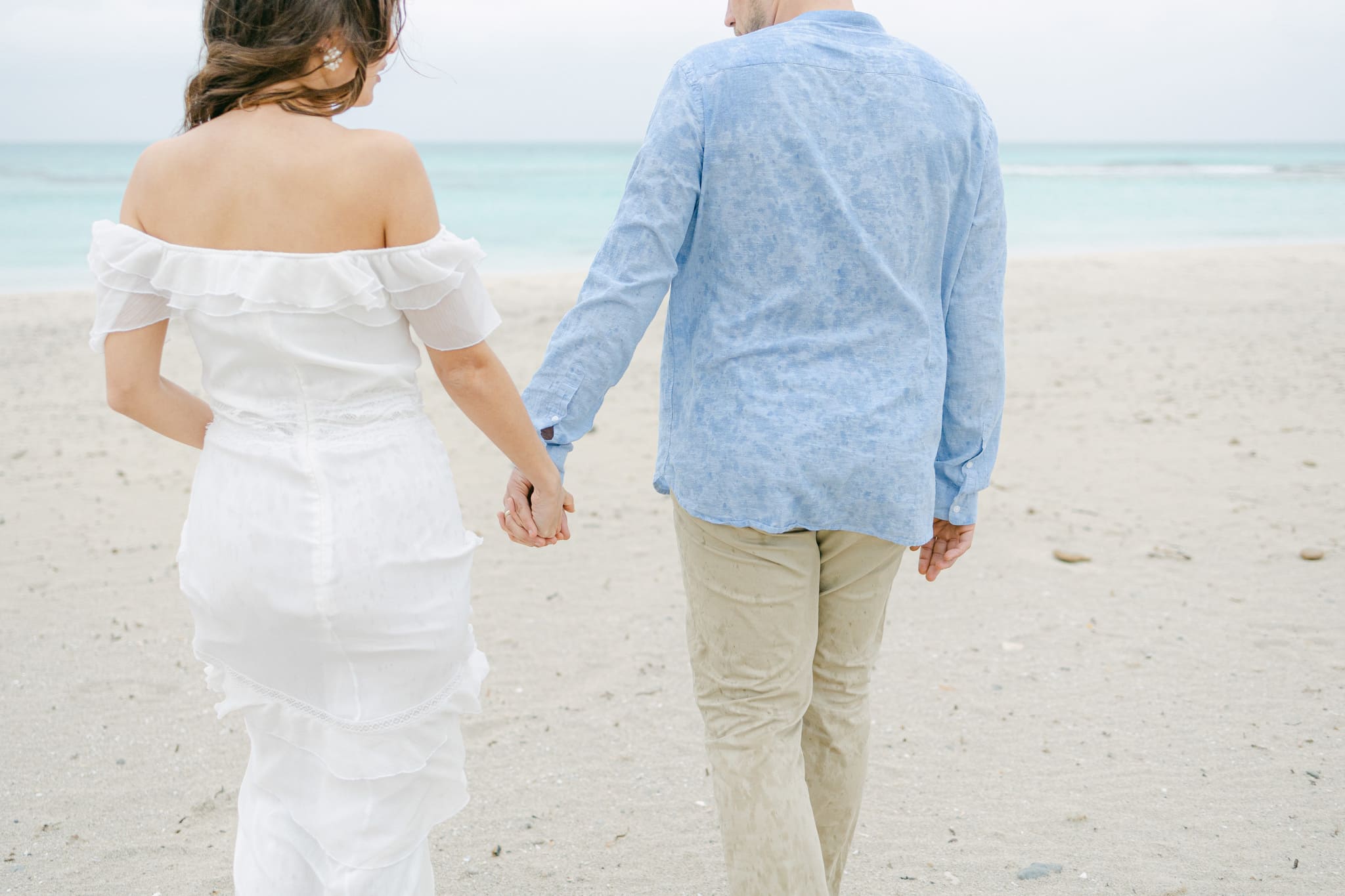 Brautpaar spaziert Hand in Hand am Strand, die Braut in einem weißen, schulterfreien Kleid und der Bräutigam in einem leichten blauen Hemd und beigen Hosen, mit Blick auf das ruhige Meer im Hintergrund.