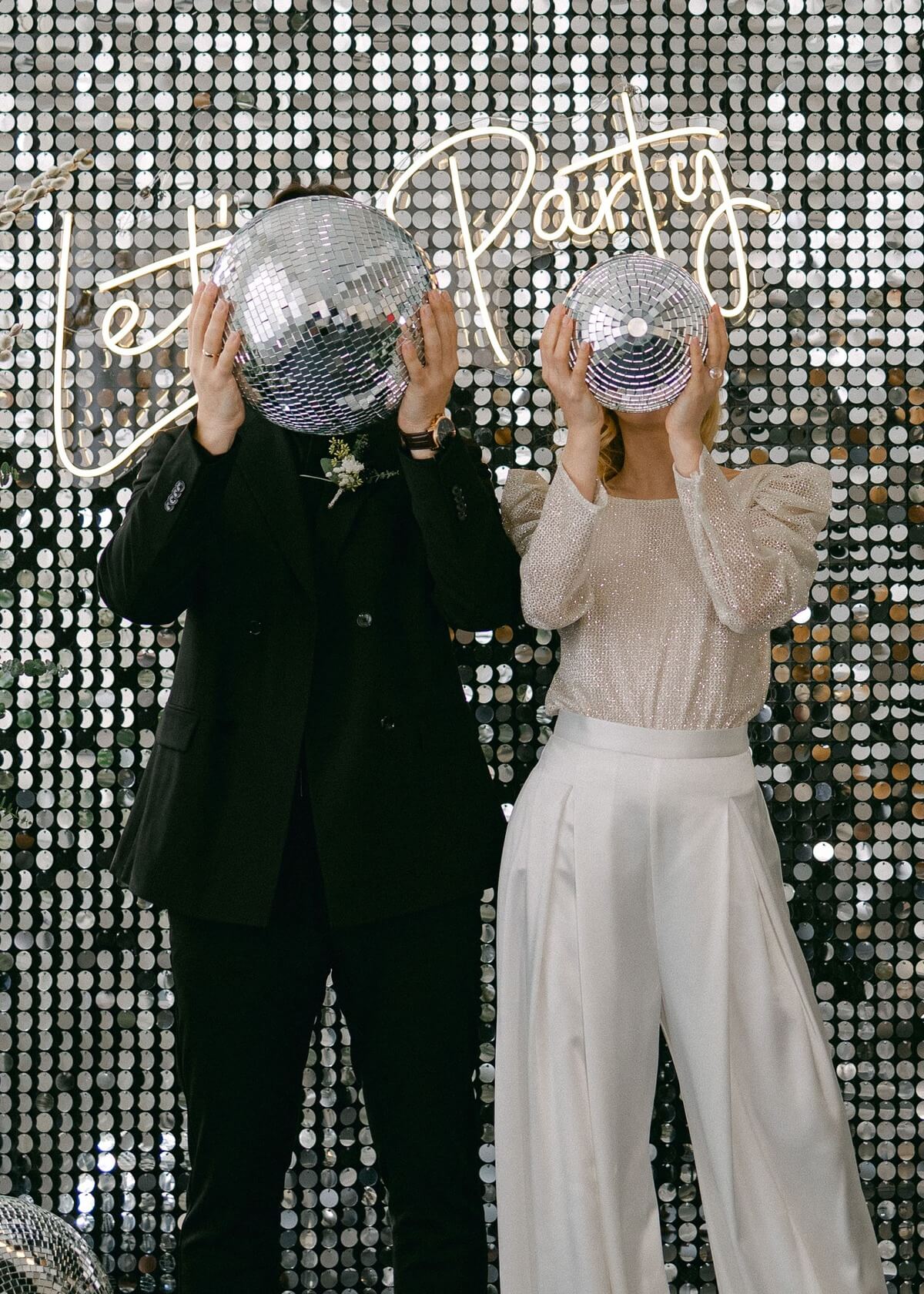 Braut und Bräutigam halten Disco-Kugeln vor ihre Gesichter bei einer stilvollen Hochzeitsparty mit einer glitzernden Kulisse und einem 'Let's Party' Neon-Schild