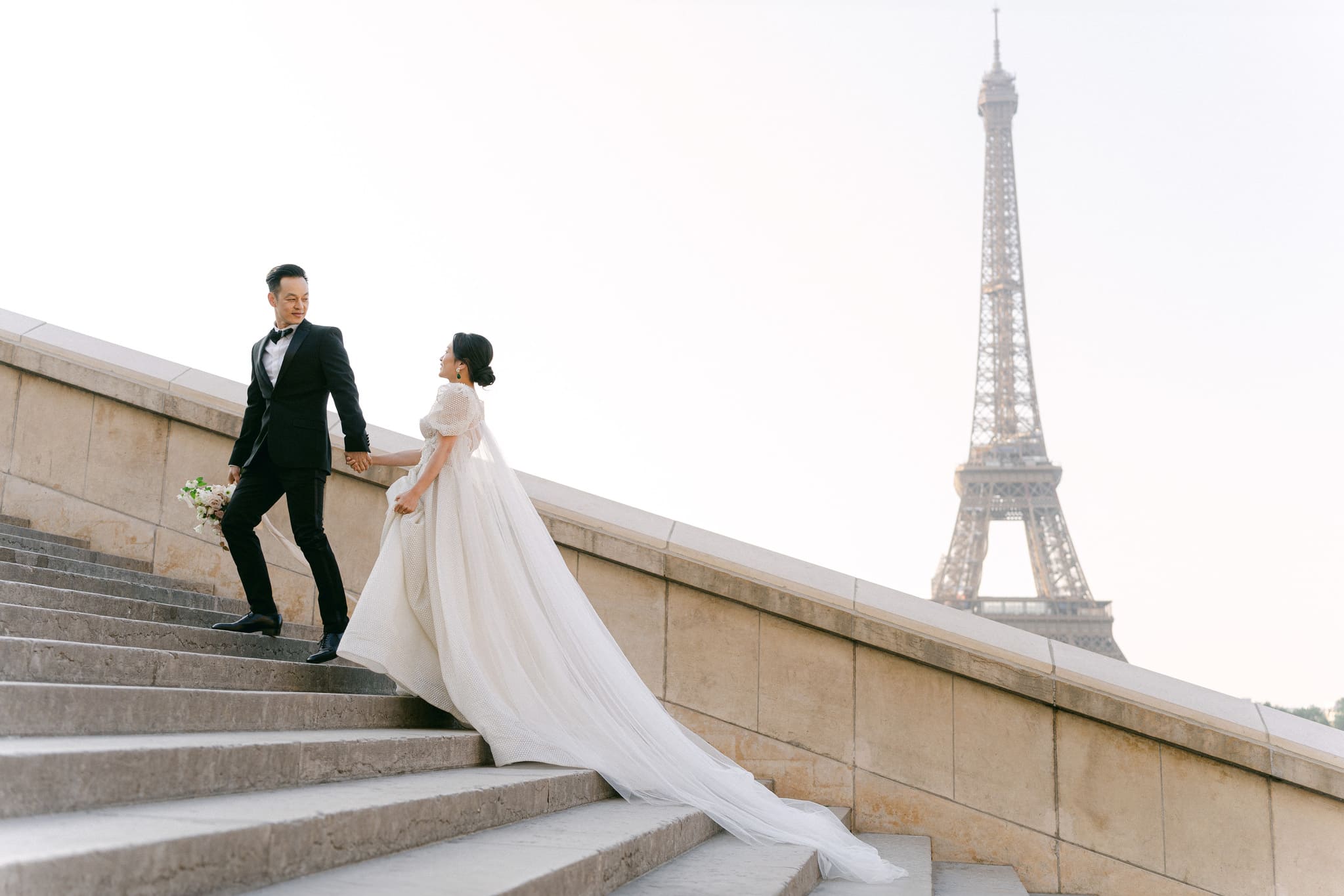 Brautpaar auf den Stufen nahe dem Eiffelturm in Paris, der Bräutigam in einem klassischen schwarzen Smoking, führt die Braut, die ein langes, elegantes Kleid mit Schleppe trägt.