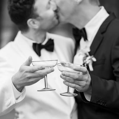 Two grooms in elegant tuxedos share a kiss and toast with martini glasses, captured in an atmospheric black and white photo.