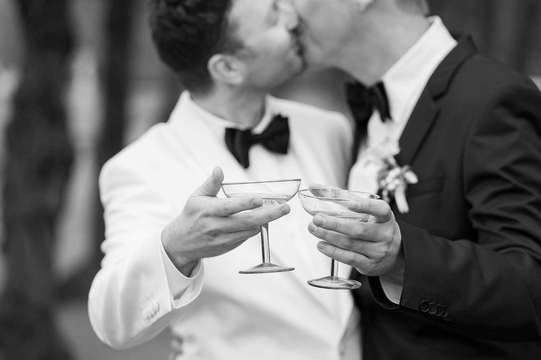 Zwei Bräutigame stoßen auf ihrer Hochzeit mit Martini-Gläsern an, während sie sich einen liebevollen Kuss geben, festgehalten in einem stilvollen Schwarz-Weiß-Foto.