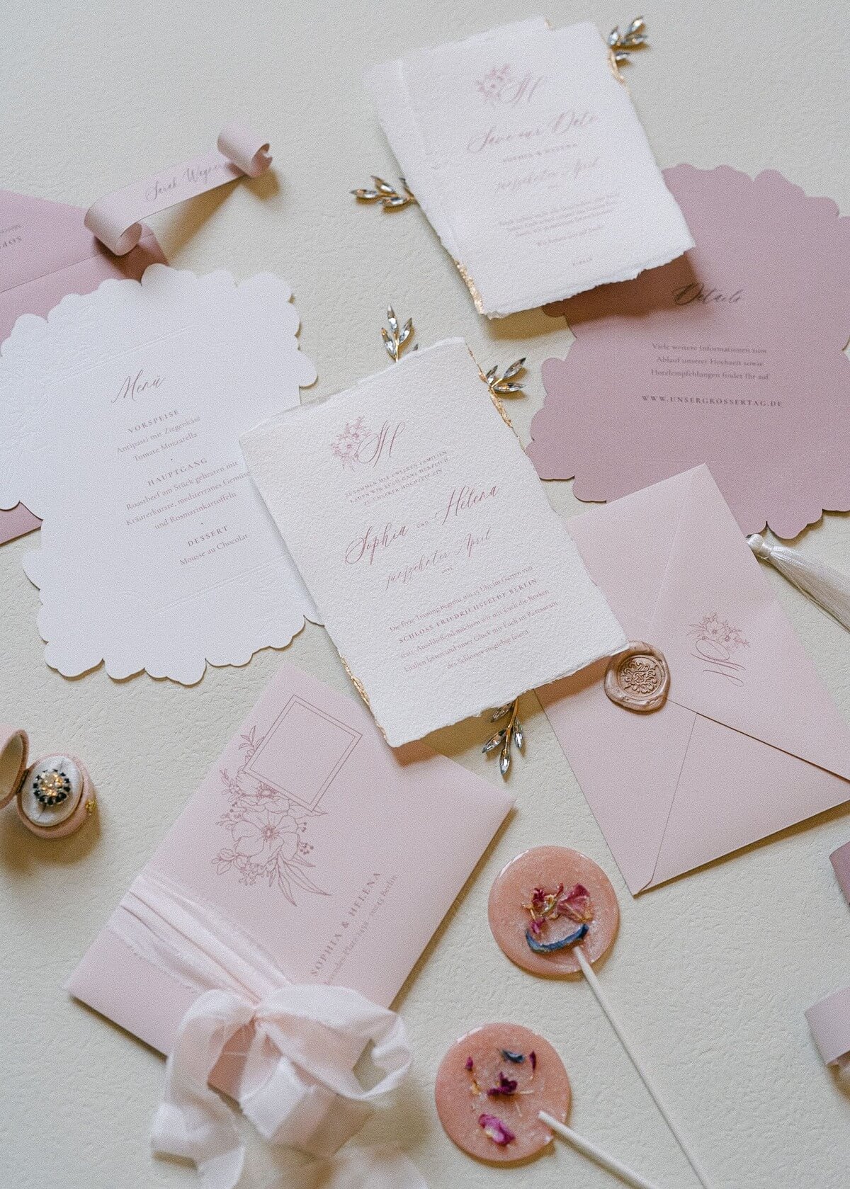 Fein gestaltetes Flatlay von Hochzeitseinladungen und Menükarten in Pastelltönen mit goldener Siegelwachs-Versiegelung und floralen Akzenten auf einem hellen Untergrund bei Hochzeit im Schloss Friedrichsfelde Berlin