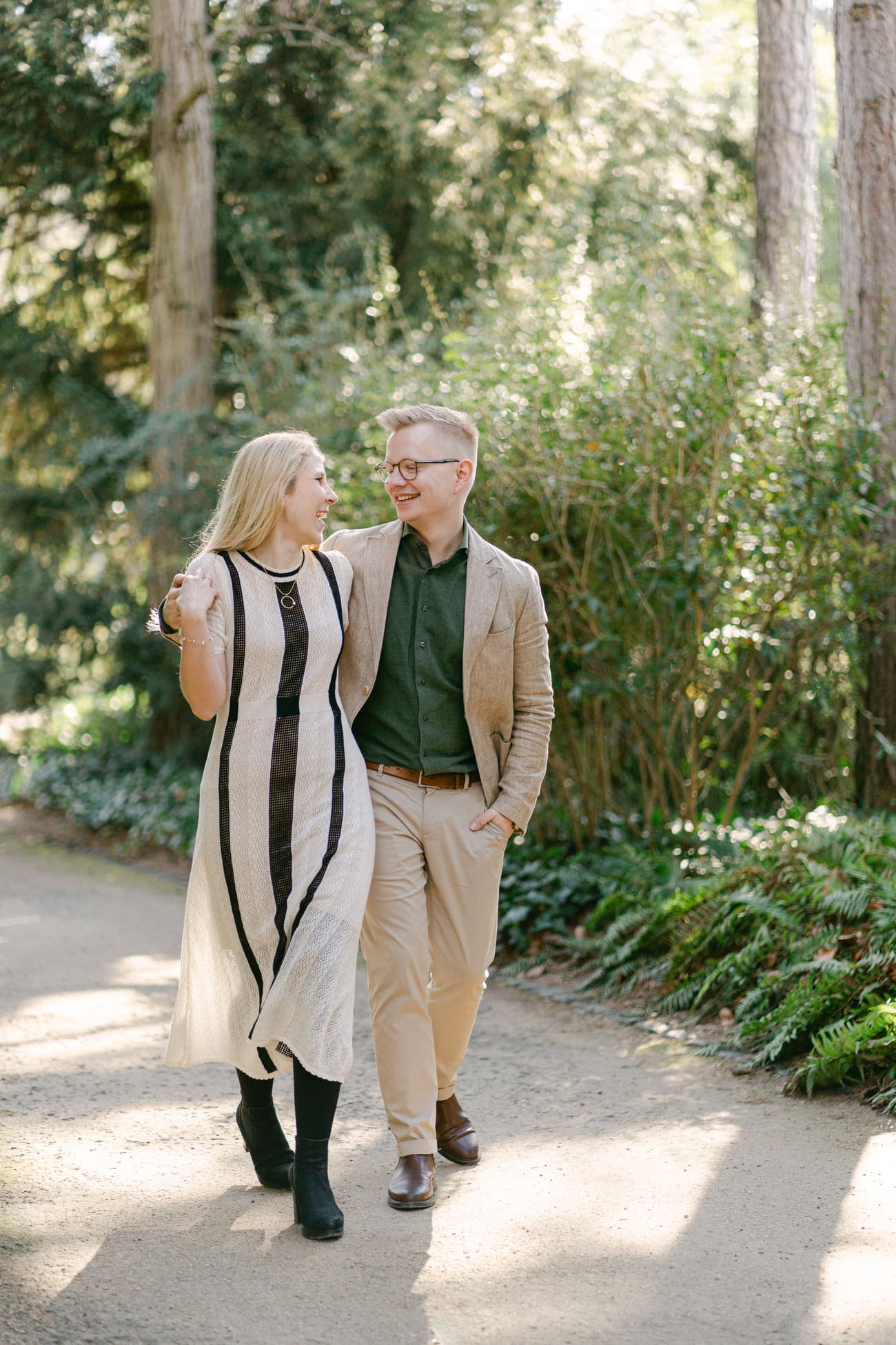 Entspannte Verlobungssession im Palmengarten: Ein glückliches Paar läuft Hand in Hand, die Frau in einem modischen, langen Kleid und der Mann in einem smarten grünen Hemd und Jackett.