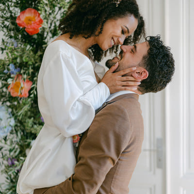 A couple in love share a tender moment during their wedding, surrounded by vibrant Bridgerton-inspired floral decorations, she in a modern wedding dress, he in a chic taupe suit.