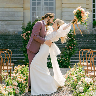 A romantic moment at a castle wedding, where the groom in an elegant burgundy suit holds his bride in a classic white wedding dress, the bride rejoicing with a large, colorful bouquet of flowers in her hand.