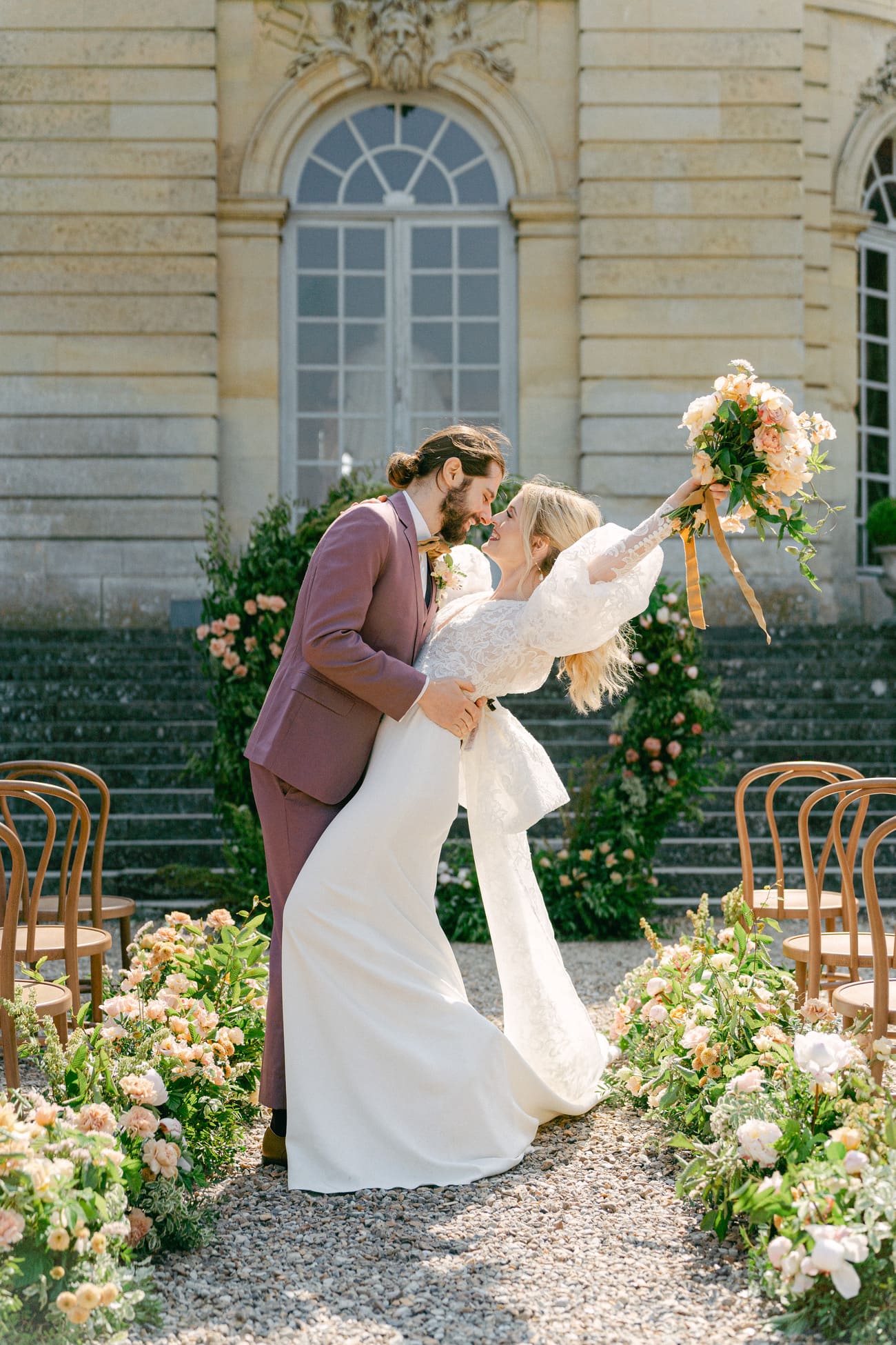 Vor dem historischen Hintergrund eines eleganten Schlosses in Frankreich nahe Paris, die Braut jubelnd mit einem großen, bunten Blumenstrauß in der Hand, gehalten von einem stilvoll gekleideten Bräutigam in einem dunkelroten Anzug.