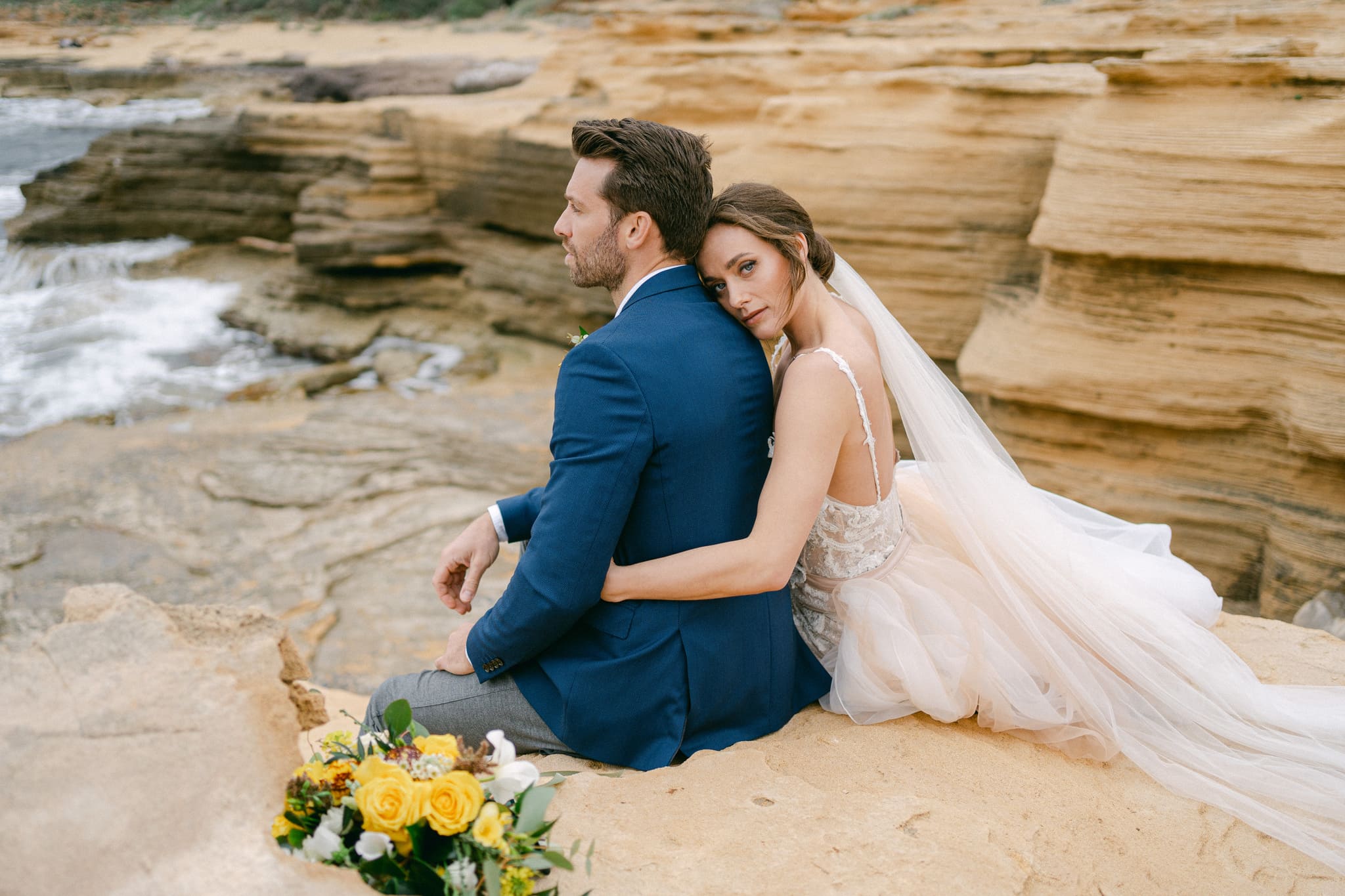 Romantische Szene eines frisch verheirateten Paares, das auf einem felsigen Strand sitzt, die Braut lehnt sich an den Bräutigam, beide betrachten die Wellen, inmitten einer atemberaubenden Küstenlandschaft.