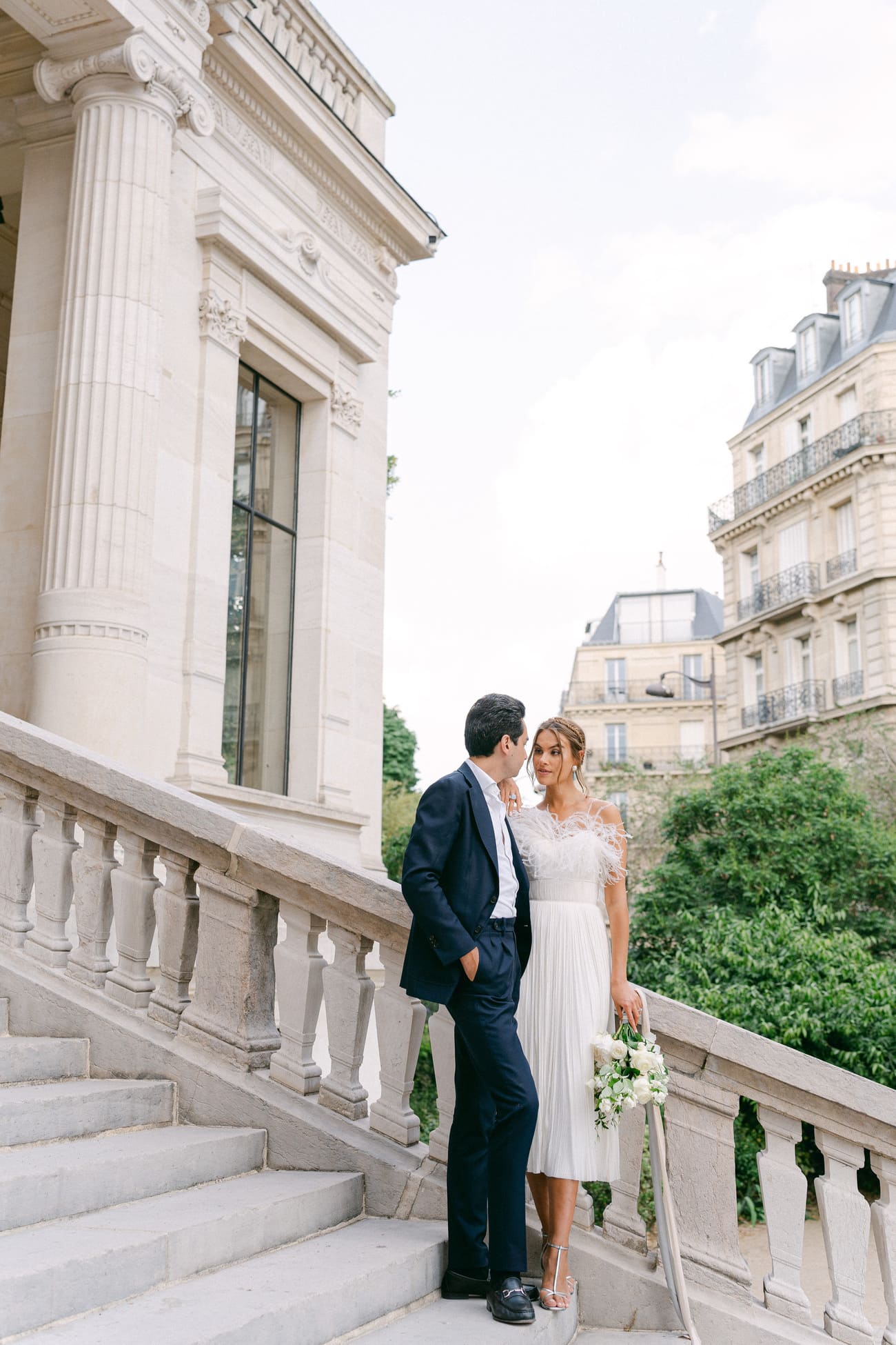 Ein Brautpaar steht an einem sonnigen Tag auf einer eleganten Steintreppe in Paris, umgeben von klassischer Architektur, die Braut in einem schulterfreien Kleid und der Bräutigam in einem dunkelblauen Anzug.