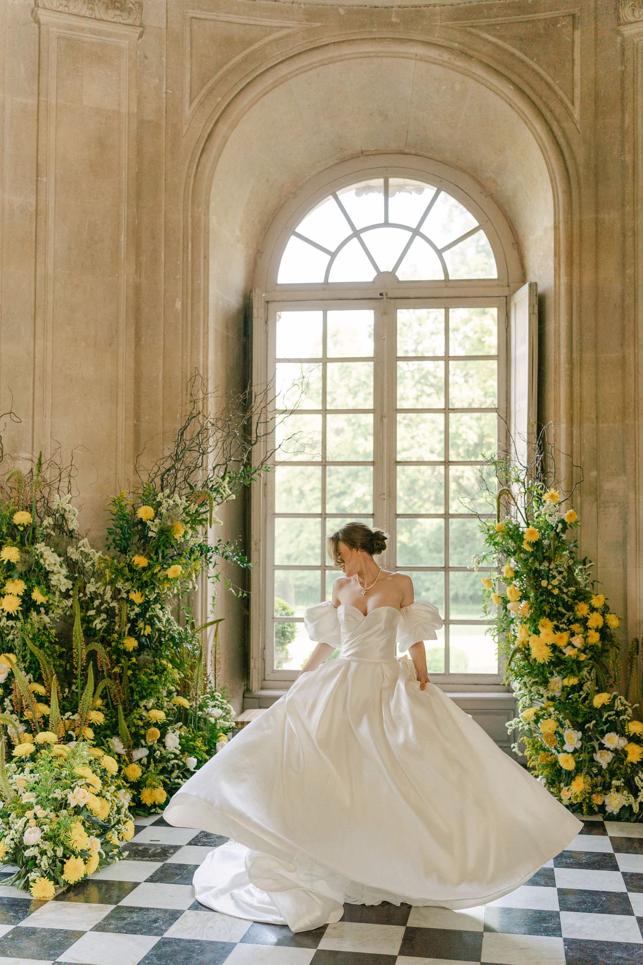 Braut in einem eleganten weißen Kleid tanzt in einem historischen Schlosssaal mit schwarz-weiß kariertem Boden, umgeben von üppigen gelben Blumenarrangements und einem großen Fenster im Hintergrund.