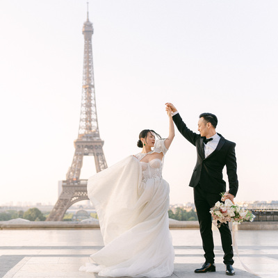 Brautpaar tanzt vor dem Eiffelturm in Paris, die Braut in einem atemberaubenden weißen Kleid mit wehender Schleppe, der Bräutigam in einem eleganten schwarzen Smoking.