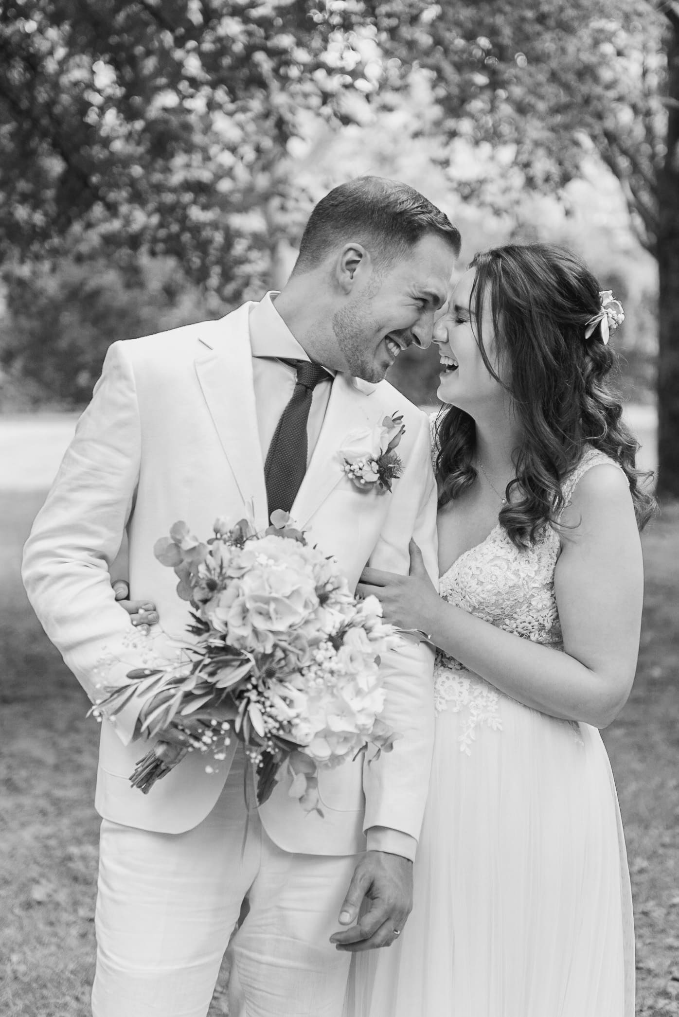 Ein glückliches Brautpaar lacht ausgelassen auf einer sommerlichen Wiese, der Bräutigam in einem weißen Anzug und die Braut in einem zarten, mit Spitze verzierten Kleid, umarmt und hält einen großen Blumenstrauß.