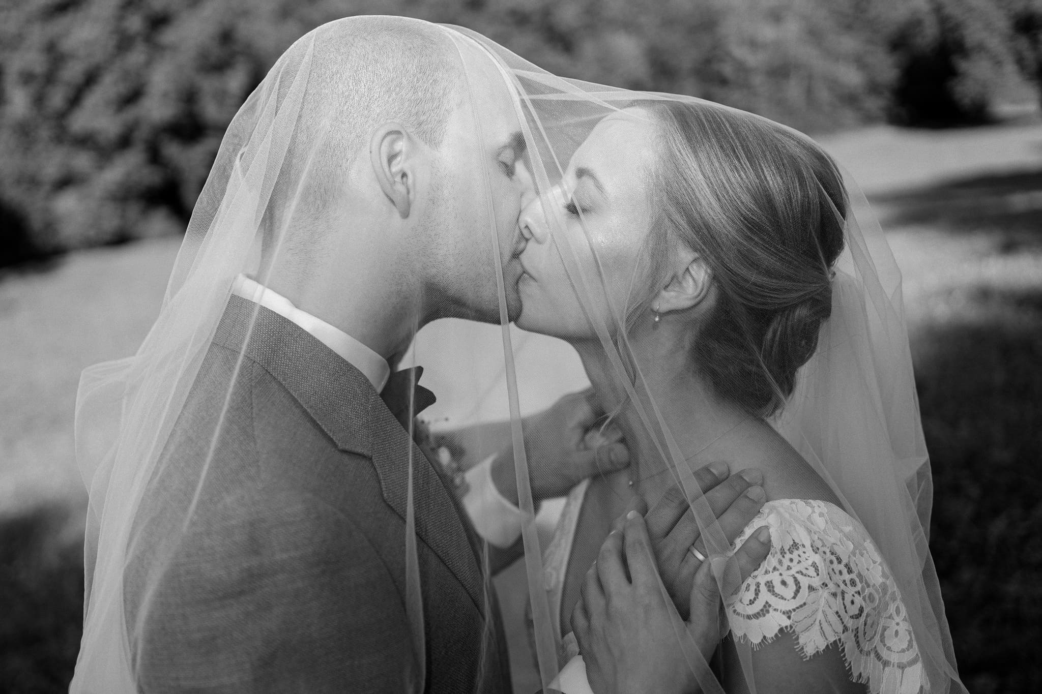 Ein emotionales Hochzeitsfoto in Schwarz-Weiß zeigt einen Bräutigam und eine Braut, die sich einen sanften Kuss geben, eingehüllt in den Schleier der Braut mit einer weichen, natürlichen Landschaft im Hintergrund.