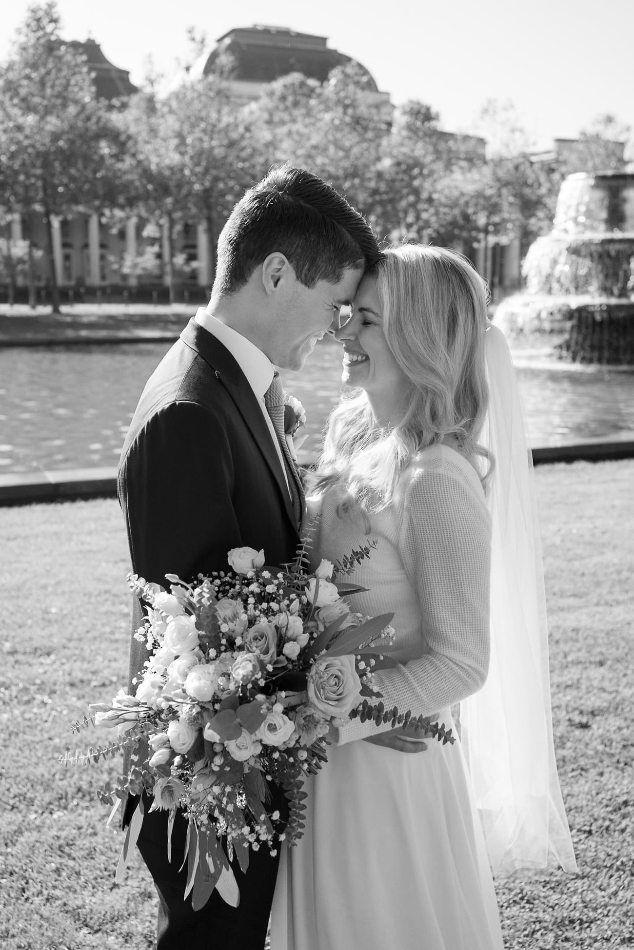Schwarz-weiß Foto von einem lächelnden Brautpaar im Sonnenschein, die Braut mit einem großen Blumenstrauß und beide lächeln sich glücklich an.